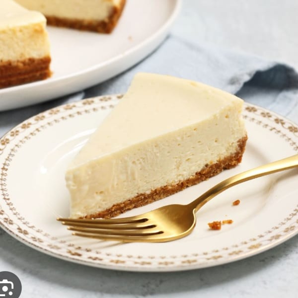 A slice of cheesecake with a golden fork on a decorative white plate, with another slice in the background.
